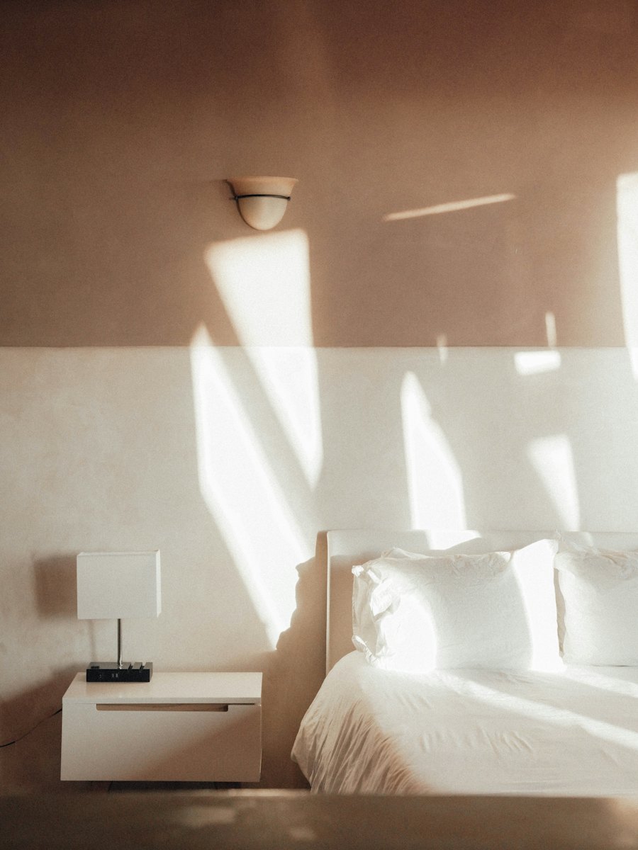 A calm bedroom with a neatly made white bed and a small nightstand in the sun